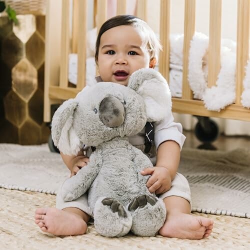 Baby sitting with a stuffed koala toy near a crib.